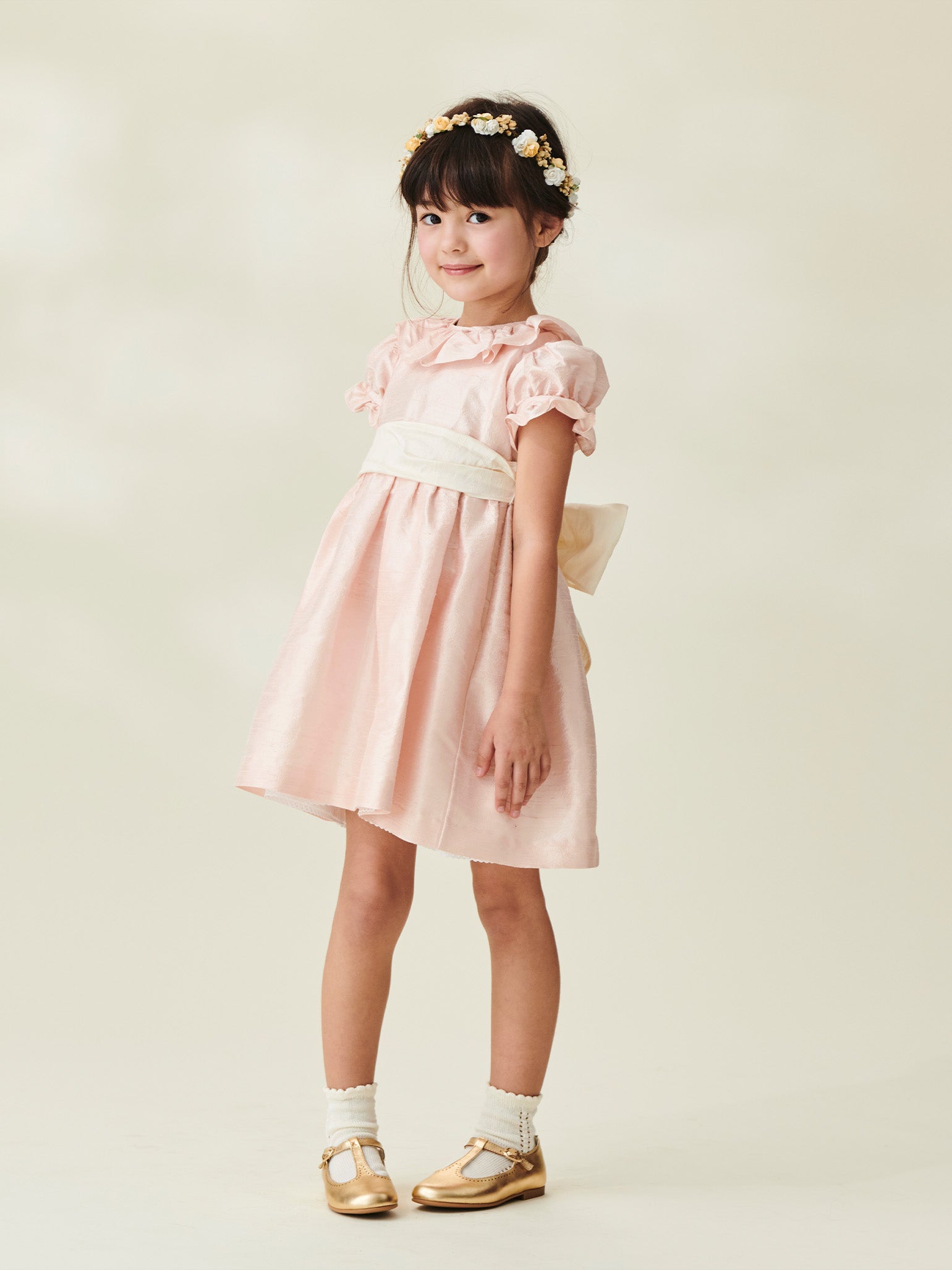 A young girl wears a charming pink satin dress with puffed sleeves and a cream sash, adorned with a flower crown and gold shoes.